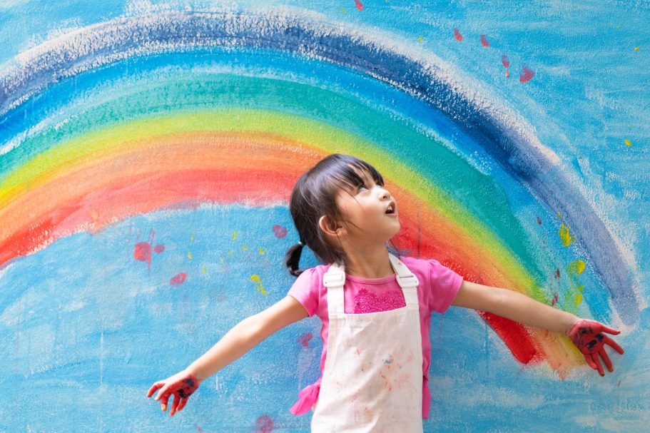 Épisode 1: Origines et perspectives petite fille en salopette blanche debout devant un mur arc en ciel qui regarde vers le ciel.